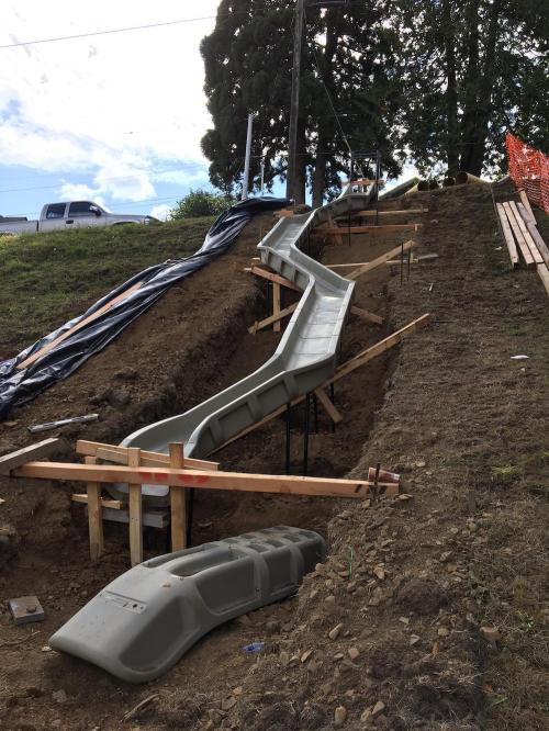 A partially constructed slide on a hillside, supported by wooden frames and surrounded by dirt and construction materials.