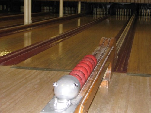 A close-up of a bowling ball return system with a red, cylindrical roller on a wooden lane.