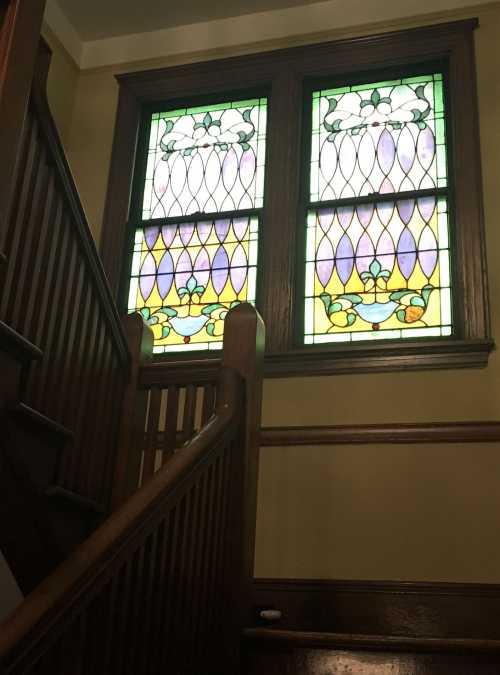 Stained glass windows with floral designs above a wooden staircase in a well-lit interior.