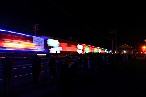 A brightly lit train passes by at night, with a crowd of people watching along the tracks.