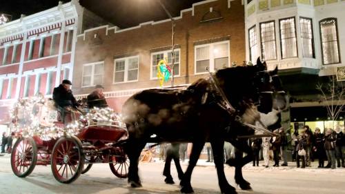 A horse-drawn carriage adorned with lights moves through a festive street, surrounded by onlookers and decorated buildings.