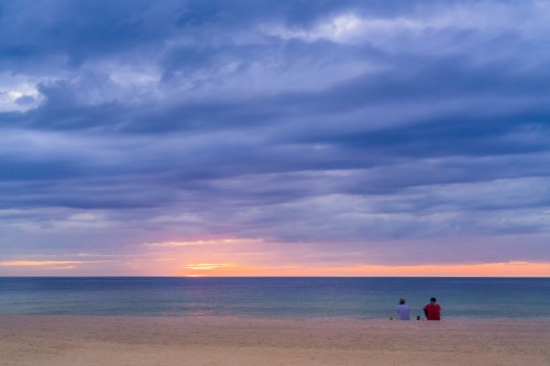 Two people sit on a beach, watching a colorful sunset over calm waters under a cloudy sky.