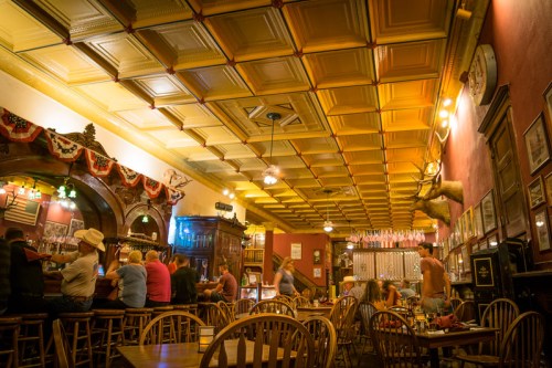 A cozy bar interior with wooden chairs, a patterned ceiling, and patrons enjoying drinks in a warm, inviting atmosphere.
