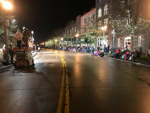 A nighttime street lined with people waiting for a parade, with festive lights on buildings and a lift in the foreground.