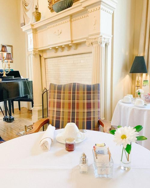 A cozy dining setup with a plaid chair, a table set for tea, and a small vase with a flower near a fireplace.