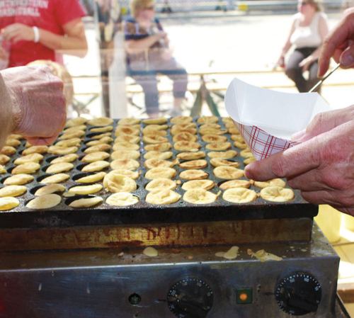Hands preparing small, round pancakes on a griddle, with a paper container ready for serving. People sit in the background.