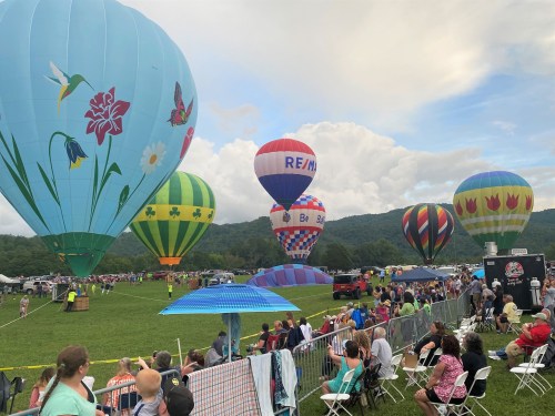 Colorful hot air balloons fill the sky above a crowd at a festival, set against a backdrop of green hills and clouds.