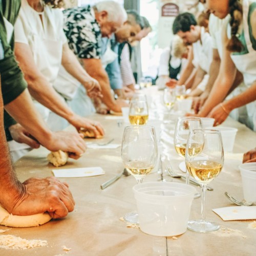 A group of people kneading dough at a long table, with glasses of wine and bowls in front of them.