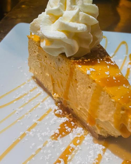 A slice of creamy cheesecake topped with whipped cream and drizzled with caramel sauce on a white plate.
