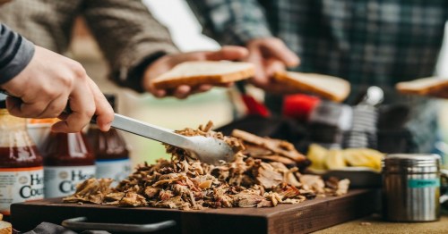 A close-up of pulled pork being served on a wooden platter, with hands reaching for bread and condiments in the background.
