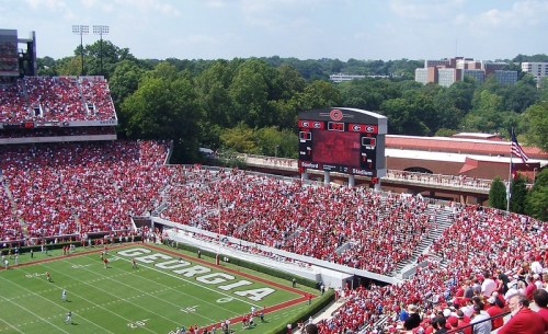A packed college football stadium in Georgia, filled with fans in red and black, with a scoreboard visible.