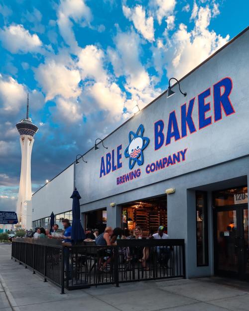 Exterior of Able Baker Brewing Company with people dining outside, featuring a tall tower in the background and a cloudy sky.