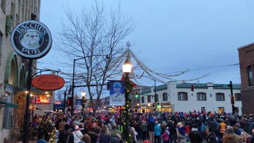 A large crowd gathers in a festive downtown area, with holiday decorations and storefronts visible.