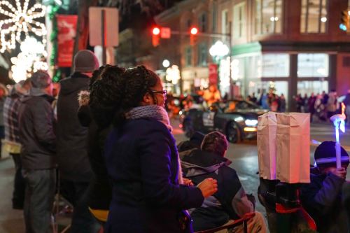 A crowd watches a festive parade at night, with colorful lights and decorations illuminating the street.