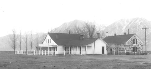 A historic farmhouse and stone building with mountains in the background, surrounded by a grassy field.