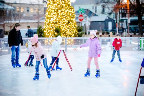 Children ice skating on a rink decorated with a glowing Christmas tree in the background.