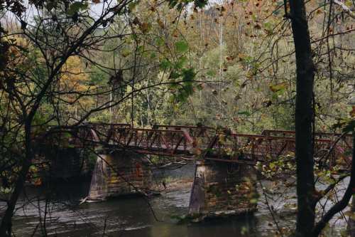 A rusted bridge spans a calm river, surrounded by trees with autumn foliage.