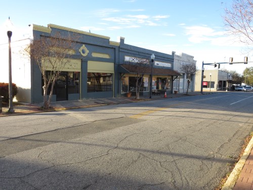 A quiet street scene featuring storefronts, trees, and a clear blue sky.