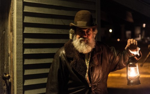 A bearded man in a brown hat and coat holds a lantern, standing near a wooden wall at night.