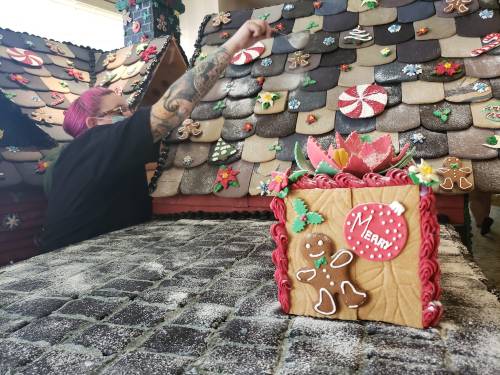 A person decorates a gingerbread house with colorful icing and candy, with a festive gingerbread box in the foreground.
