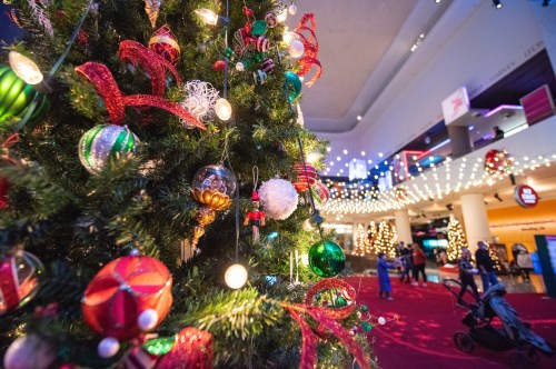 A close-up of a decorated Christmas tree with colorful ornaments, set in a festive indoor space with lights.