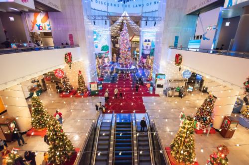 A festive atrium decorated with Christmas trees, lights, and holiday displays, bustling with visitors.