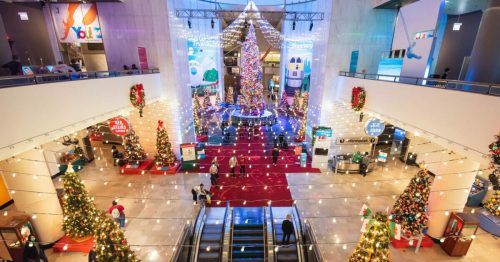 A festive atrium decorated with Christmas trees, lights, and holiday displays, bustling with visitors.