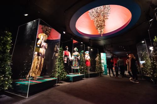 Exhibition space featuring colorful costumes in glass displays, surrounded by festive decorations and visitors.