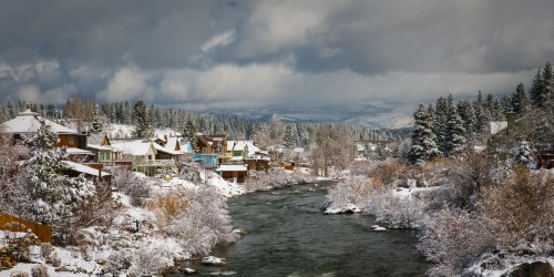 A snowy landscape with charming houses along a river, surrounded by trees and mountains under a cloudy sky.