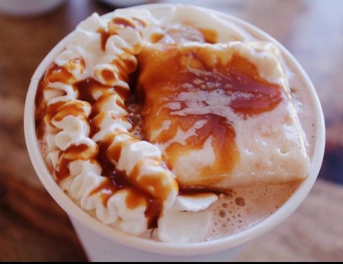 A close-up of a cup of hot chocolate topped with whipped cream and a caramel drizzle, featuring a pastry on top.