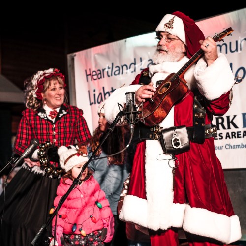 A man in a Santa costume plays a ukulele, while a woman in a festive dress and a child look on, smiling.