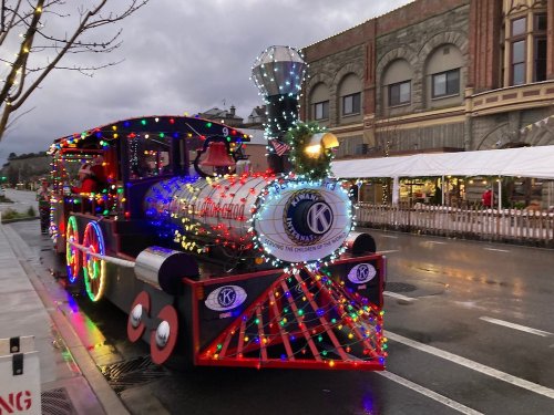 A brightly lit, festive train decorated with colorful lights parked on a street, with buildings in the background.