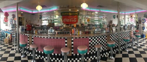 A retro diner interior featuring a checkered floor, a long counter with stools, and colorful neon lights.