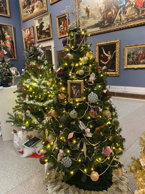 A beautifully decorated Christmas tree with lights and ornaments, surrounded by festive artwork in a gallery.