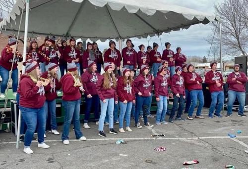 A large group of people in matching maroon hoodies singing on a stage under a tent, with a cloudy sky above.