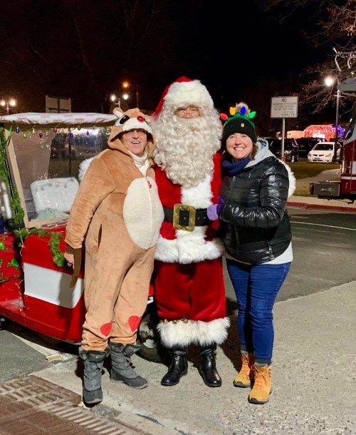 Three people pose together: one in a reindeer costume, one dressed as Santa, and one in a festive hat, with holiday lights in the background.