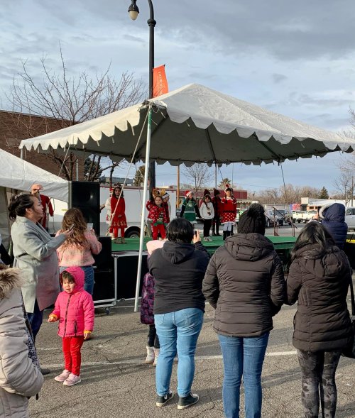 A group of performers in red costumes dance on stage under a tent, while an audience watches in a parking lot.