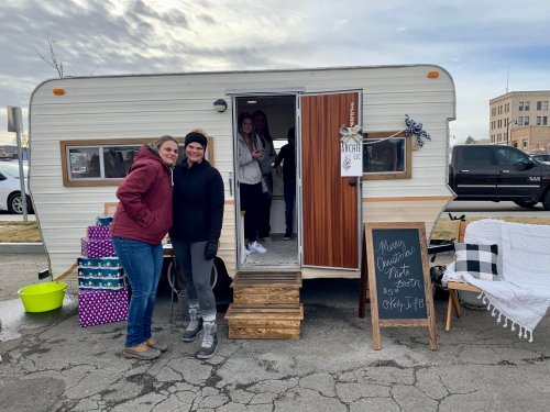 Two women stand in front of a vintage trailer, smiling, with gifts and a chalkboard sign nearby.