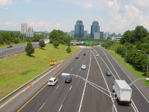 A view of a busy highway with cars, green trees, and tall buildings in the background under a blue sky.