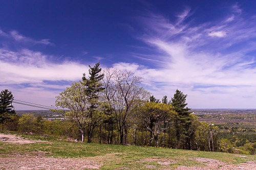A scenic view from a hilltop, featuring trees and a vast landscape under a blue sky with wispy clouds.
