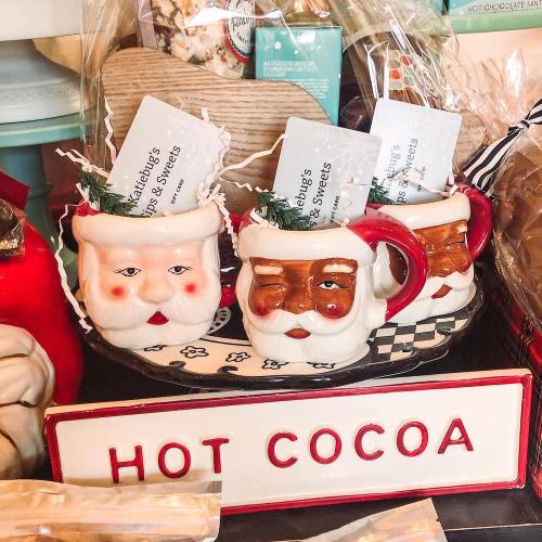 Three festive mugs shaped like Santa faces, filled with treats, displayed on a tray labeled "HOT COCOA."