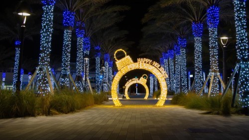A festive pathway lined with blue-lit palm trees and a glowing archway that reads "Happy Holidays."