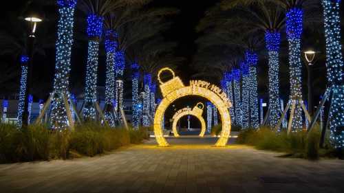 A festive pathway lined with blue-lit palm trees and a glowing archway that reads "Happy Holidays."