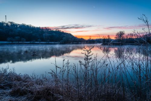 Frosty landscape at dawn, with a calm river reflecting the colorful sky and silhouetted plants in the foreground.