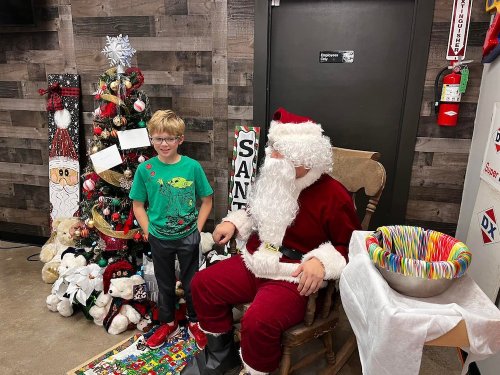 A child stands next to Santa Claus, who is seated in a chair, with a decorated Christmas tree in the background.