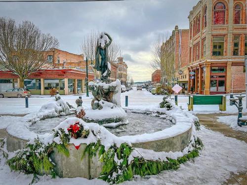 A snow-covered fountain surrounded by buildings and trees, with festive decorations in a winter scene.