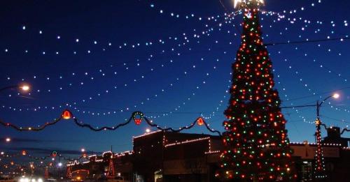 A brightly lit Christmas tree surrounded by colorful lights against a twilight sky, with festive decorations along the street.