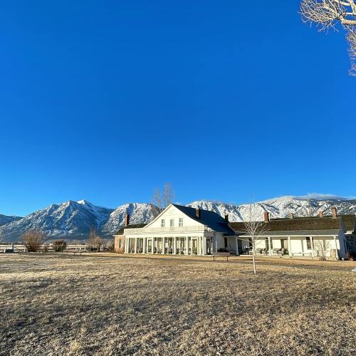 A large white house with a porch, set against snow-capped mountains and a clear blue sky.
