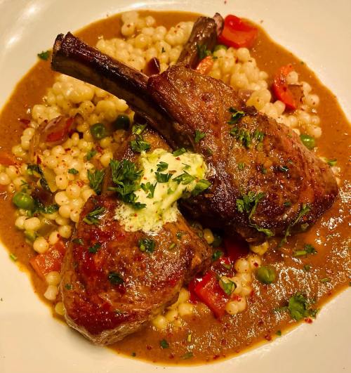 Two lamb chops on a bed of couscous with vegetables, topped with a dollop of herb butter and garnished with parsley.