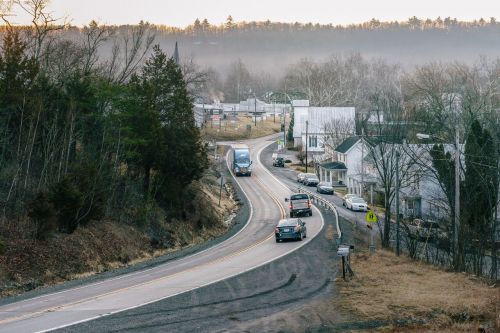 A winding road through a small town, with vehicles and buildings surrounded by trees and a misty atmosphere.